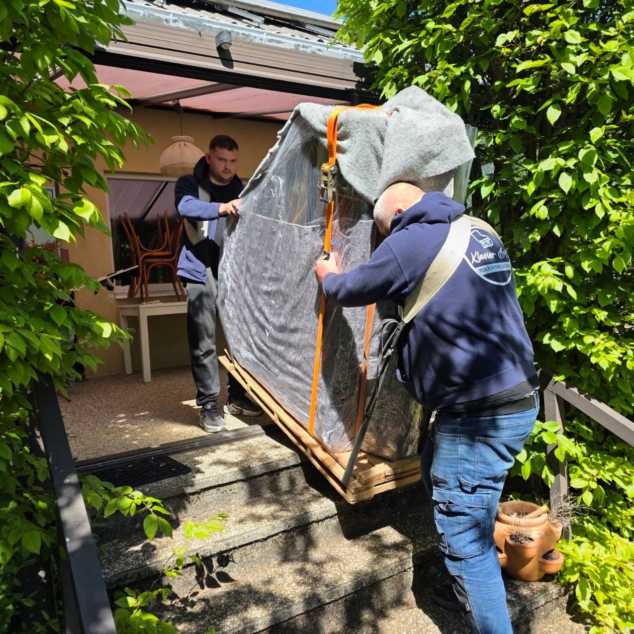 Piano transport Krems: a wrapped piano carried outdoors along a garden path to the van
