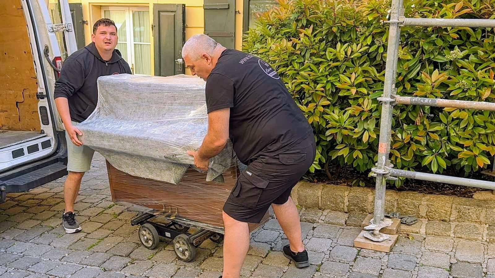 Zwei Mitarbeiter von MK Klavier Art tragen ein verpacktes Klavier auf dem Transportwagen zum Fahrzeug