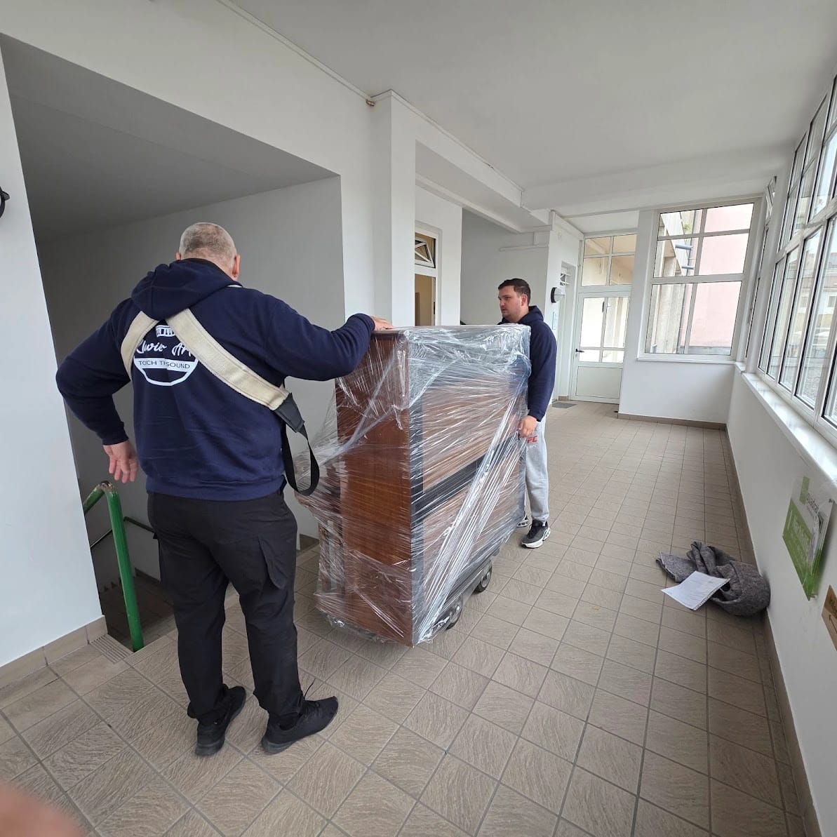 Zwei Männer tragen ein verpacktes Klavier durch einen hellen Gang