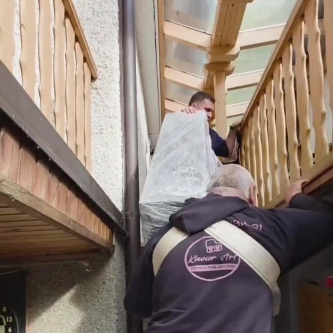 Two men carrying a wrapped piano up a covered outdoor staircase